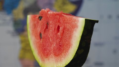 Close-up of a slice of red watermelon with seeds. The camera flies around. Parallax effect. In the background, a blurry map of the world