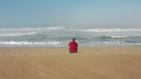 Aerial view of picturesque seascape with endless ocean in the background. Lonely man observes the ocean waves, while sitting on the seashore. High quality 4k footage