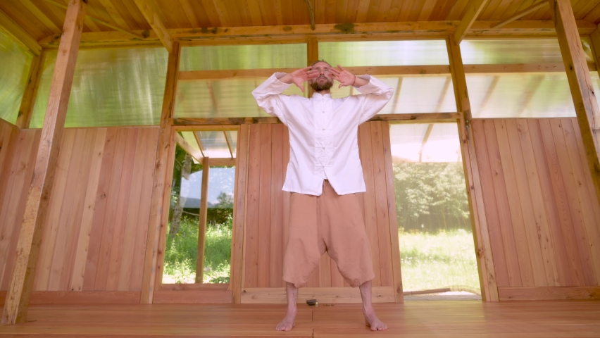 A long-haired Caucasian man in light and loose clothing practices qigong tai chi in a wooden practice room in summer. Slow movements harmony and calm