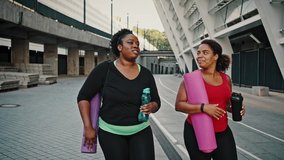 Sport and friendship. Outdoor portrait of two positive chubby african american women going home after fitness training, carrying water bottles and fit mats, slow motion - Powered by Shutterstock - Get 15% off with code: PIKWIZARD15