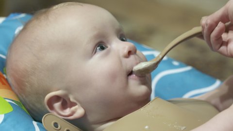 Little cute baby boy eating first food from spoon with his mother at home, mom feeding pureed food to her son. High quality 4k footage