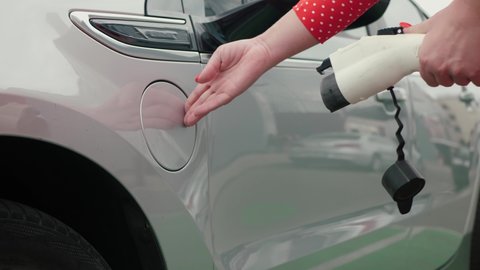 Female hands plugging in power supply cable to charge EV close up. Unrecognizable woman in red dress with white dots opens charging port lid an electric car and connects plug
