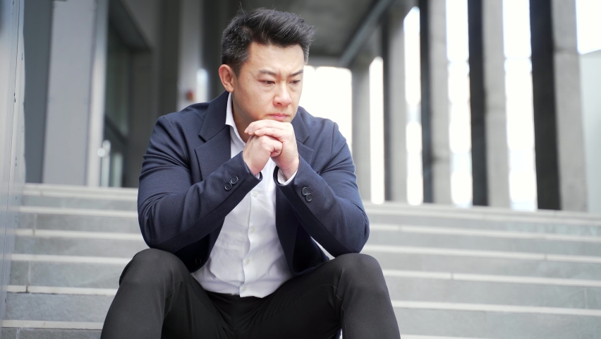 Young asian depressed man sitting on the stairs outdoors street near office business center building. Upset male businessman lost his job due financial crisis and quarantine. Businessman has problems | Shutterstock HD Video #1078984430