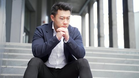 young asian depressed man sitting on the stairs outdoors street near office business center building. Upset male businessman lost his job due financial crisis and quarantine. Businessman has problems
