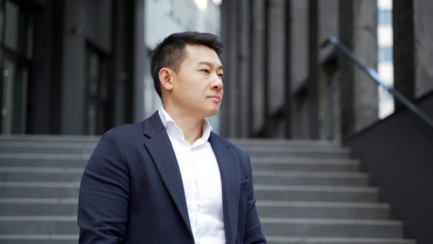 portrait of a young confident successful asian businessman looking at camera. Happy business man, close up smile standing on urban background of modern office building outdoors. Male in a formal suit Royalty-Free Stock Footage #1078984451