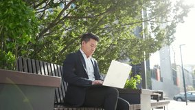 asian male entrepreneur freelancer sitting outdoors in city park a bench talking online a video call using laptop Business man office worker working on urban street background Businessman communicates