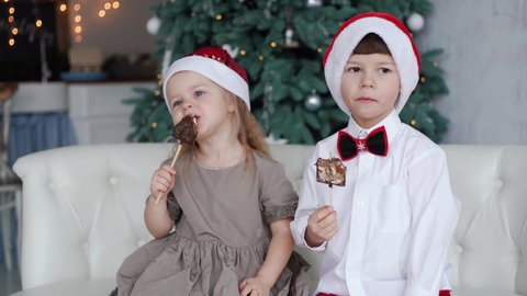 4k video portrait of two cute little children eating Christmas chocolate sweets happily. Craft chocolate handmade sweets in hands of kids. Brother and sister wearing red holiday Santa hats