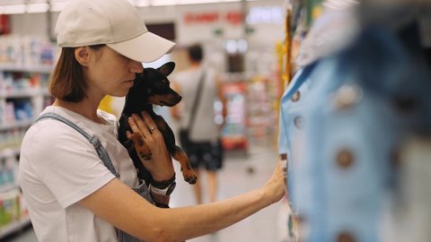 Young woman shopping at the supermarket with little toy terrier dog in her hands. She choosing dog food for her pet. High quality 4k footage