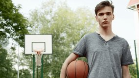Boy standing on a basketball court and holding a basketball. Cinematic shot of teenager standing with a ball in his hand and a basketball basket behind him. Video of a confident face of a basketball p