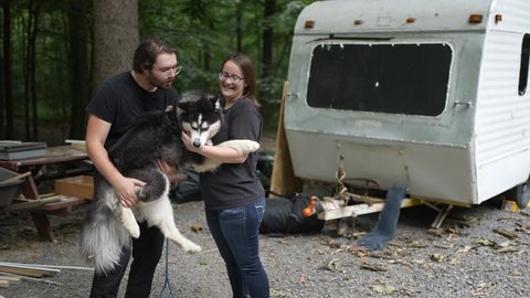 Man and woman play with Husky dog in front of customized travel trailer under construction.