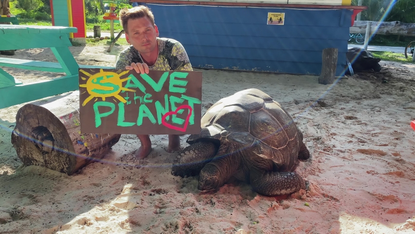 A guy with a poster in his hands that says "Save the Planet" and next to him is a huge tortoise of a rare species of which there are very few, aldabras in the Seychelles