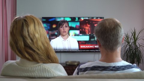 Family Couple Watching TV News Sitting on Couch in Living Room Together. TV Presenter Telling Shocking Breaking News with Scenes of Violence, Fires, Protests and Clashes. Rear View with Dolly-in
