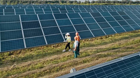 Three employees of alternative power plant walking and talking about scheme of solar panels. Technician and investor walking in Solar cell Farm through field of solar panels checking the Solar