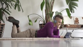 Young cute modern guy using tablet with headphones to have online videocall conversation with friends or family on the floor at home
