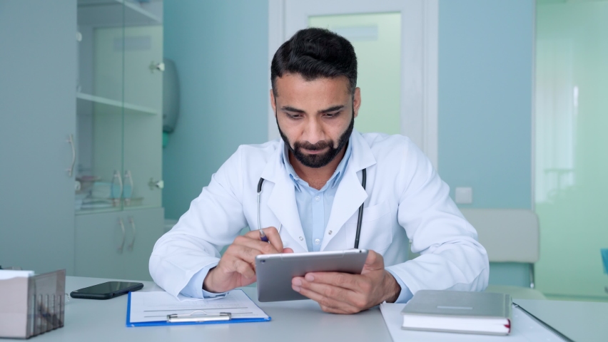 Indian male serious general practice doctor therapist in modern clinic in white coat using tablet device, typing texting consulting remotely, writing health data. Telemedicine health care concept. Royalty-Free Stock Footage #1078998617