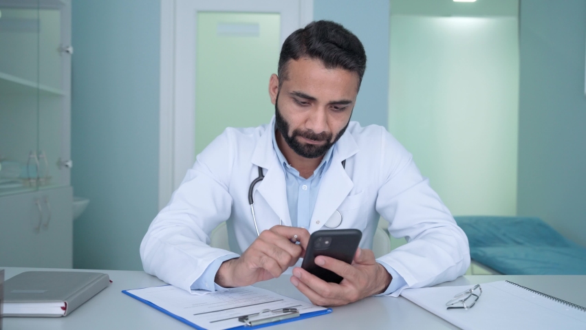 Indian male doctor therapist in modern clinic wearing white coat uniform using cell mobile smartphone apps, texting and writing health data. Medicine technologies health care concept. Royalty-Free Stock Footage #1078998620