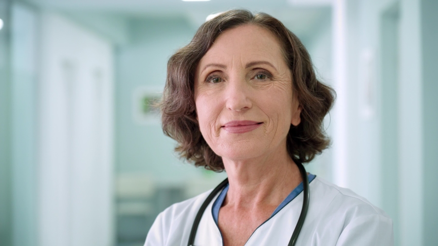 Portrait of happy smiling older senior female professional doctor physician pediatrician nurse wearing white robe standing in modern private clinic hospital looking at camera. Close up. Royalty-Free Stock Footage #1078998623