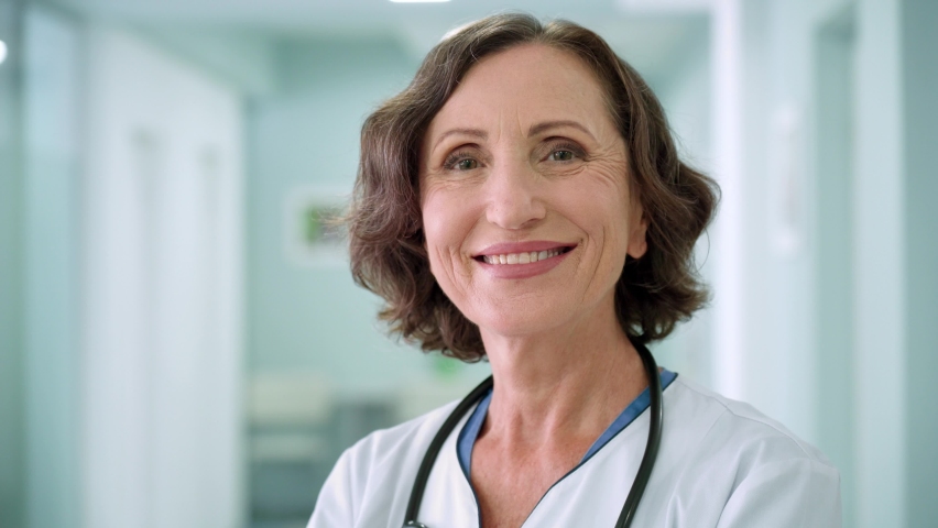 Portrait of happy smiling older senior female professional doctor physician pediatrician nurse wearing white robe standing in modern private clinic hospital looking at camera. Close up. Royalty-Free Stock Footage #1078998623