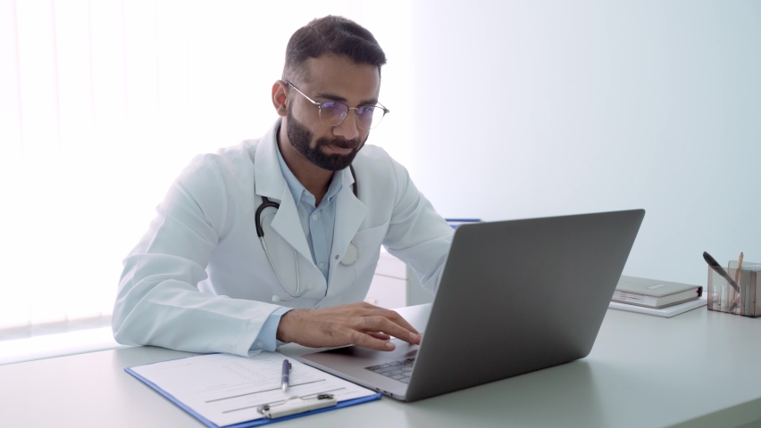 Indian male serious doctor medical worker in modern clinic wearing eyeglasses and white doctor's coat typing using laptop computer writing health personal data. Telemedicine healthcare concept. Royalty-Free Stock Footage #1078998626