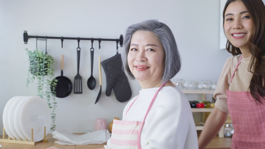 Portrait of Asian lovely family, young daughter stand with old mother. Attractive female and senior elderly mom wear apron enjoy activity at home to bake croissant in kitchen and smile, look at camera Royalty-Free Stock Footage #1079007446