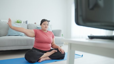 Asian large oversize women doing yoga workout in living room in house. Overweight sport girl sit on floor exercise by streching hand, follow instruction from online trainer to lose weight for health.