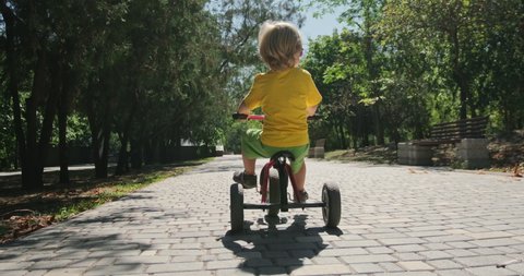 Strong long haired toddler in colorful clothes rides tricycle along empty paved road in green park on sunny day backside view