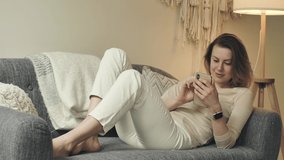 Pretty female person sits on cozy sofa at her apartment with smartphone in hands. Woman with bare feet scrolls and browses, texts messages. Charming girl types on display while lying on comfy couch.