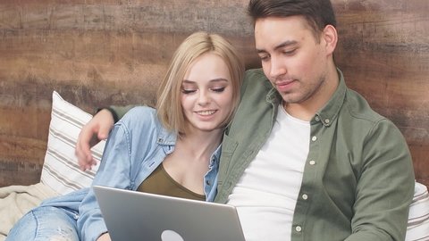 Young couple watches movie on laptop lying on bed in their bedroom.