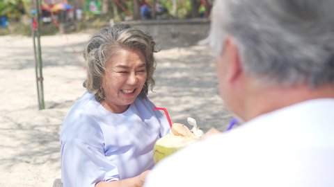 Asian family senior couple sitting on blanket have picnic drinking coconut juice together on the beach. Healthy elderly husband and wife relax and enjoy outdoor lifestyle activity on summer vacation