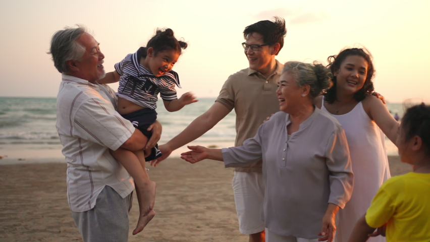 4K Happy big Asian family on beach travel holiday vacation. Multi-generation family enjoy and fun walking together on the beach at summer sunset. Grandparents embracing and carrying grandchild girl.
