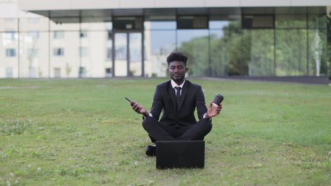 Young African American businessman in black formal suit, relaxing in the lotus position, sitting on green meadow in front of modern office building. Yoga practice for office workers