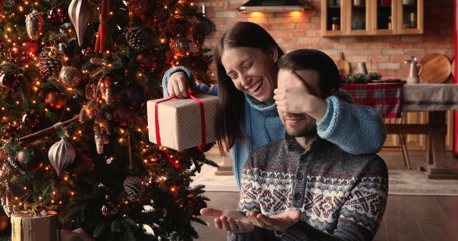 Smiling young 30s woman covering eyes of smiling curious husband, giving wrapped box with Christmas gift, sitting together near decorated festive tree, New Year winter holidays family celebration. Royalty-Free Stock Footage #1079025230