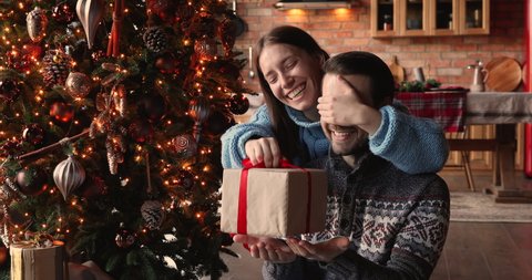 Smiling young 30s woman covering eyes of smiling curious husband, giving wrapped box with Christmas gift, sitting together near decorated festive tree, New Year winter holidays family celebration.