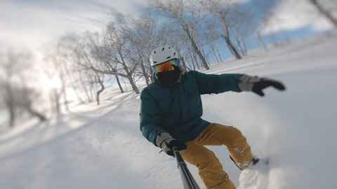 Guy doing snow splash rising on magnificent sunny day. Concept of extreme, sport, winter, freeride, snowboarding. Man riding on snowboard with selfie stick in his hand between trees on slow motion.