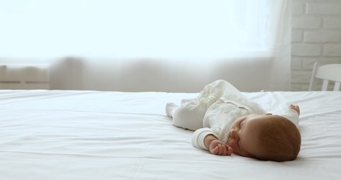 Serene newborn fell asleep alone on bed. Sweet peaceful baby in white bodysuit lying on bedsheet enjoy daytime nap, 0-6 child sleep in domestic room. Healthcare, paediatric, carefree babyhood concept