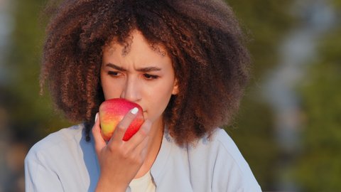 Close up Afro american woman african curly girl sitting outdoors sniffs red fresh ripe apple delicious fruit feeling frustrated loss of smell symptom coronavirus disease covid virus looking at camera
