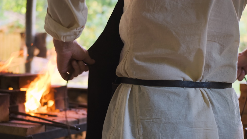 blacksmith in medieval village in outdoor forge prepares to work and ties his leather apron at back. In background, furnace is already lighting up to heat steel.