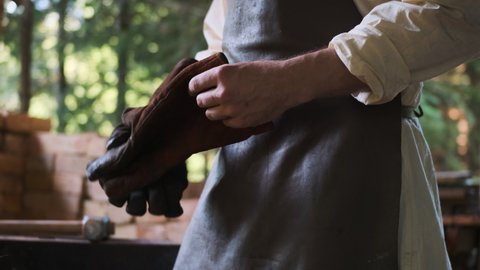 smithy worker puts on fireproof gloves before working with red-hot metal. medieval blacksmith shop on street, where simple hard workers peasants in leather apron and white shirt work.