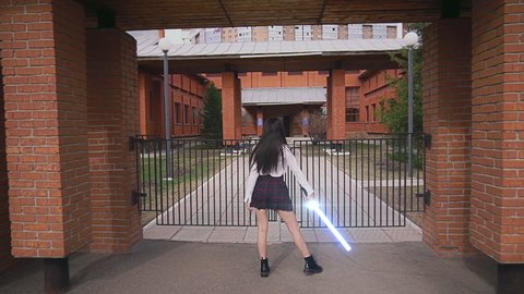 Modern female knight standing with lightsaber in front of building on sunny day. Posing woman warrior with light saber sword. Girl in Scott pattern skirt and white shirt holds laser blade in hands.