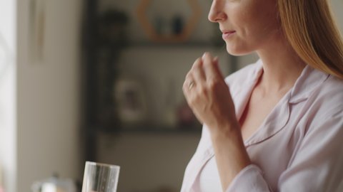 Tilt up shot of beautiful pregnant woman taking medication from pillbox and drinking glass of water while standing in kitchen at home