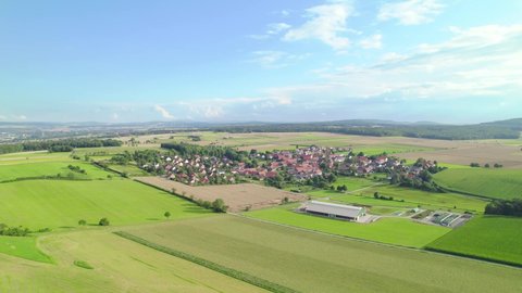 Aerial view of a metall cow shed next to a beautiful German village surrounded by green meadows and summer landscape  Tilting and panning drone shot 