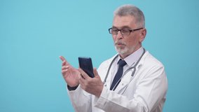 Doctor speaks by video link on the phone, standing in studio on a blue background. Adult doctor in a medical gown and glasses communicates via video link through the phone.