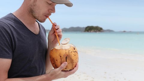Close-up view of man drinking coconut on white sand beach against tropical island. Real time in 4K resolution. 