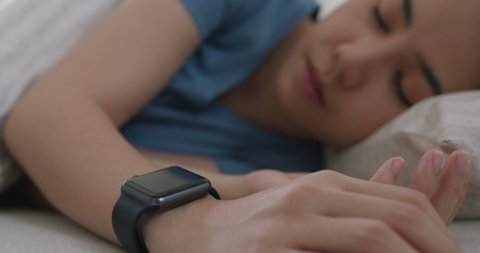 Asia teen woman wear smartwatch for sugar tracker, blood pressure tracking, resting sleep rate on arm IoT tech collect data app device relax body on cozy bed in future life at home.