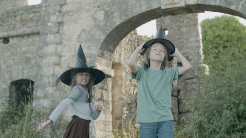 Boy and girl standing in witch hats among stone walls of castle. Happy Caucasian kids having fun together, pretending to be magicians, conjuring, speaking incantations. Witchcraft concept