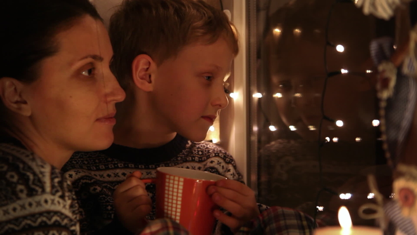 Mother and son looking out window awaiting Santa with Christmas lights in background. Holidays, magic, Christmas concept.