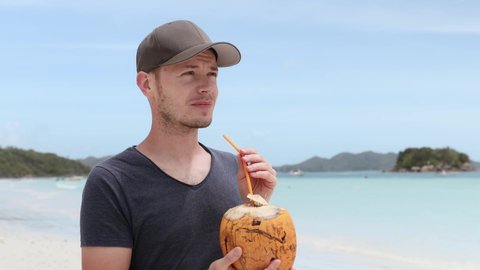 Man drinking coconut on white sand beach against tropical island. Real time in 4K resolution. 
