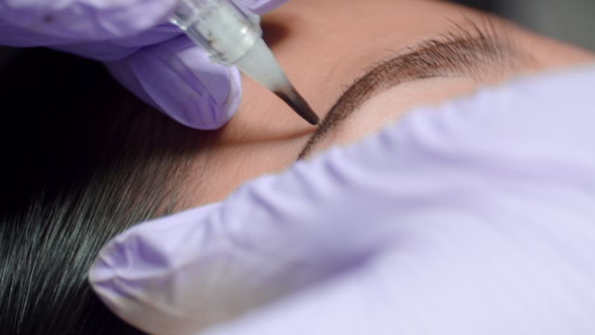 Close up microblading eyebrow tattoo. Beautician in gloves makes permanent makeup correction of the shape of the eyebrows to a young women. Eyebrow tattoo in a beauty salon. | Shutterstock HD Video #1079070020
