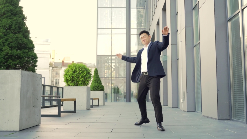 Cheerful happy asian business man dancing walk the corridor backdrop a modern office building, outside, outdoors Funny successful businessman in suit rejoices joyfully jump celebrates victory, success | Shutterstock HD Video #1079071583