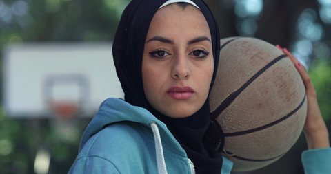 Cinematic close up shot of young happy arab woman wearing burqa is smiling satisfied with her results in camera on basketball outdoor court background after finishing a friendly game match.